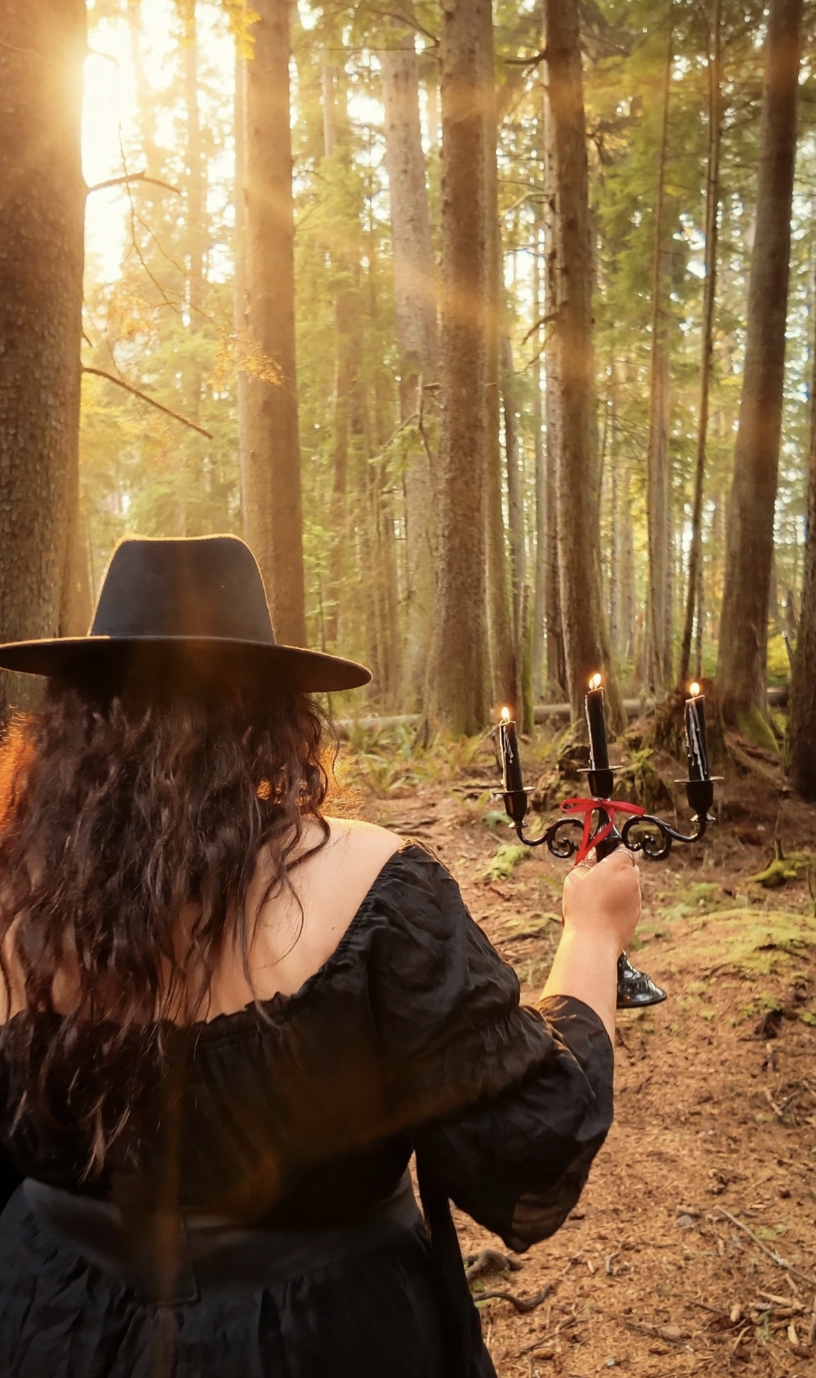 A woman in a black dress and wide-brim hat carrying a candelabra through a sunlit forest
