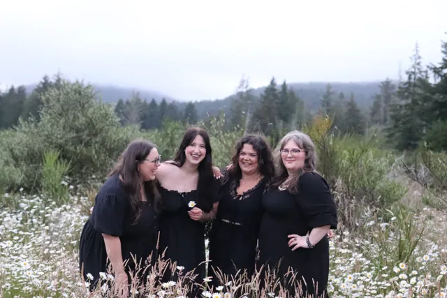 Four women in black dresses laughing together in a daisy field